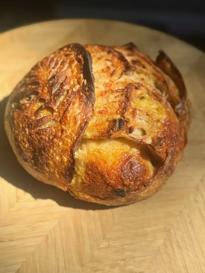 Round sourdough loaf with a deep score on a wooden board in direct sunlight