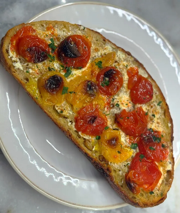 Slice of toasted sourdough topped with ricotta and blistered cherry tomatoes on a white plate