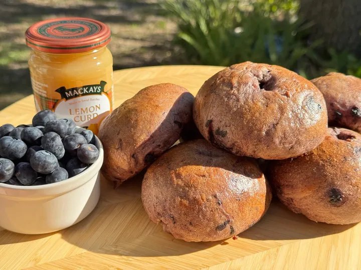 Blueberry sourdough bagels on a wooden board beside fresh blueberries and a jar of lemon curd