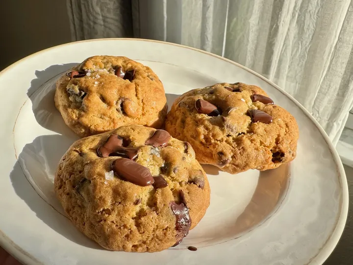 Three chocolate chip cookies topped with flaky sea salt on a cream-colored plate in sunlight