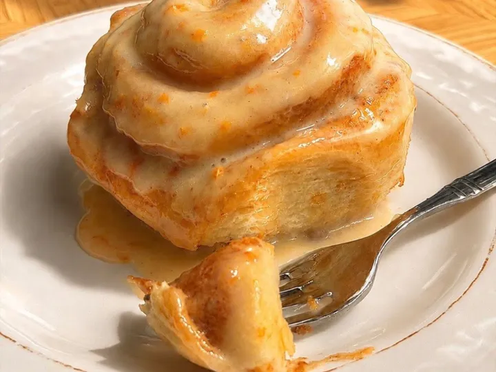 Orange sweet roll with glossy citrus glaze on a white plate beside a fork