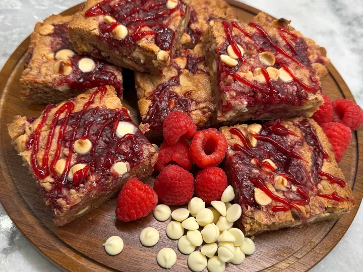 Raspberry white chocolate blondie bars on a wooden plate with fresh raspberries and white chocolate chips