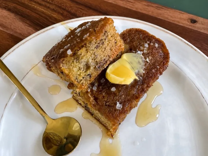 Sliced salted maple banana bread on a white plate with butter, syrup, flaky salt, and a gold spoon